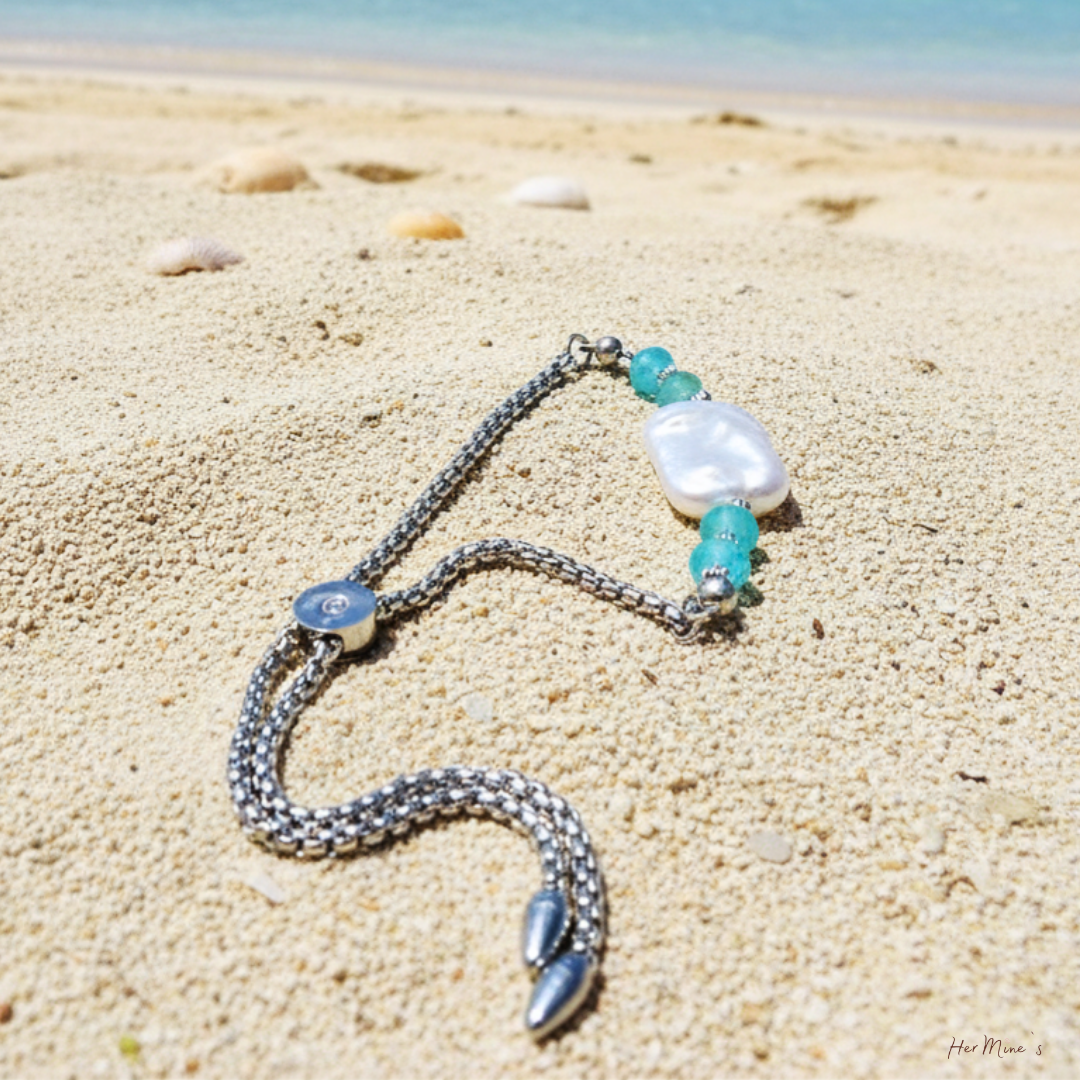 LAGOON armband från HerMine's liggandes på sand vid havet — blå topaz, barock keshipärla och boxkedja i rostfritt stål