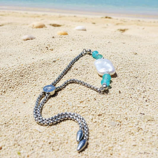 LAGOON armband från HerMine's liggandes på sand vid havet — blå topaz, barock keshipärla och boxkedja i rostfritt stål