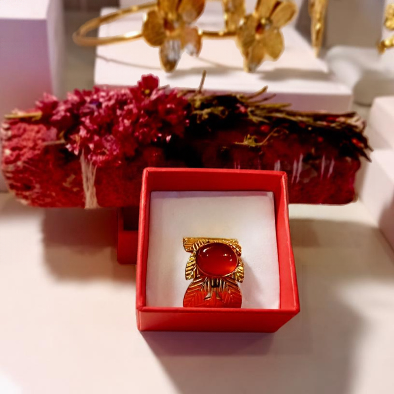 MEROPE-ring i öppen röd smyckeask, guldfärgade blad med röd karneolcabochon, torkade blommor i bakgrunden