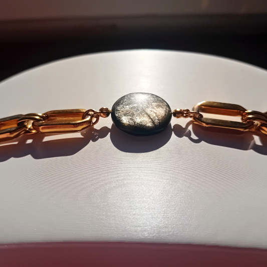 Armband med en stor, rund, golden mica sten i mitten, infattad mellan guldpläterade länkar i paperclip-stil, på ljus bakgrund i solljus