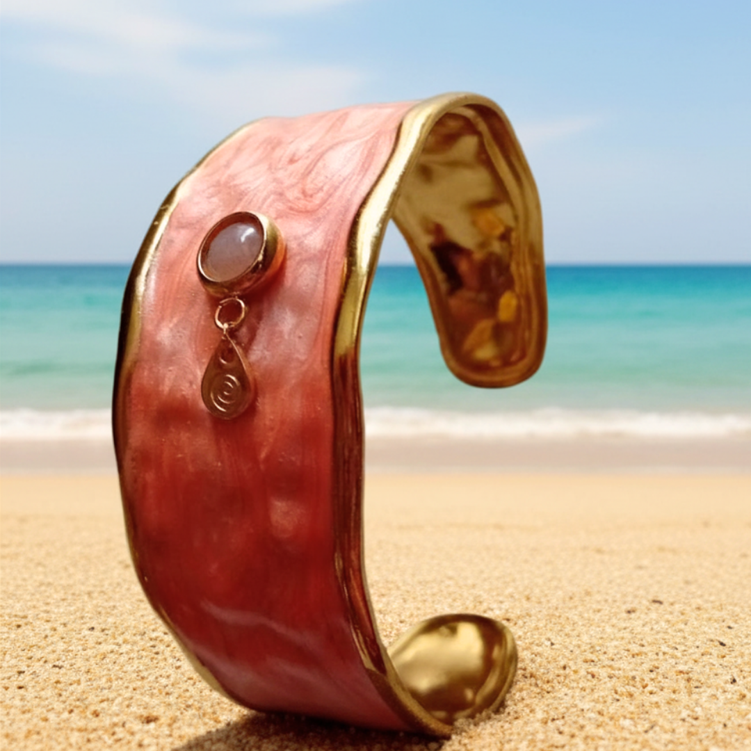 SUNKISSED manschettarmband från HerMine's – bred korallrosa emaljmanschett med solsten och guldspiral, stående på sidan i strandsand med turkost hav i bakgrunden
