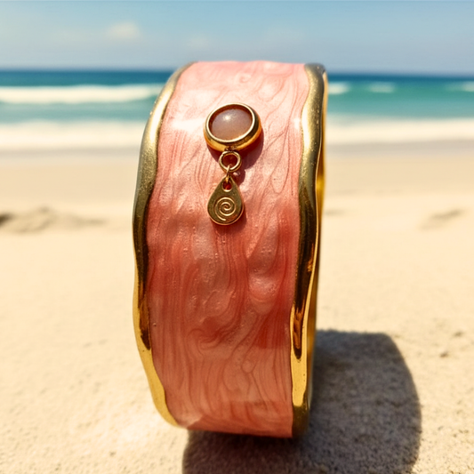 Brett manschettarmband i korallrosa emalj med guldkanter och solsten, stående i strandsand – SUNKISSED av HerMine's