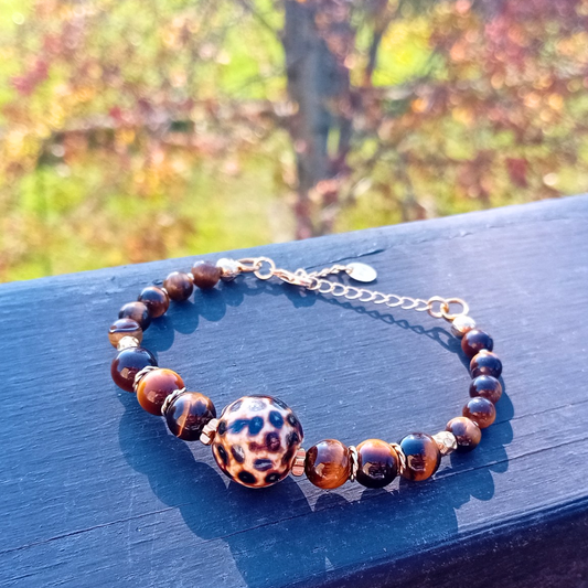 ZAHARA-armband på mörkt trä, runda tigerögonpärlor och en leopardmönstrad keramikpärla med guldpläterad förlängningskedja, höstlöv i bakgrunden