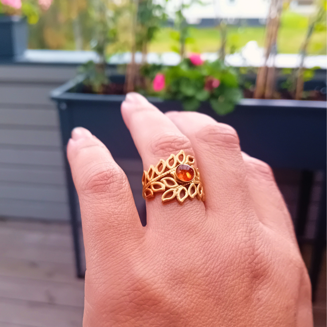 ARDEN-ring med cognacfärgad bärnsten och bladkrans i guld på fingret, med rosa blommor i bakgrunden