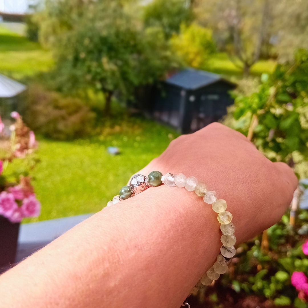 LIVIA-armband på handleden med fasetterade, ljusa gröna prehnitpärlor, mörkgröna serafinitpärlor och en hamrad stålkula, trädgård i bakgrunden