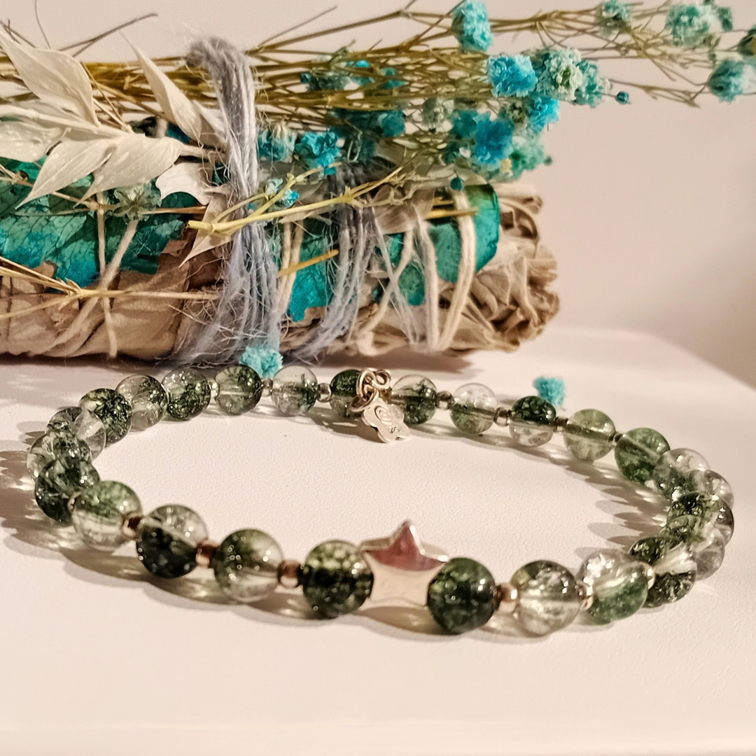 MIRA-armband på vit yta med gröna fantomkvartspärlor och en silverstjärna, en rökelsebunt med torkade blommor i bakgrunden