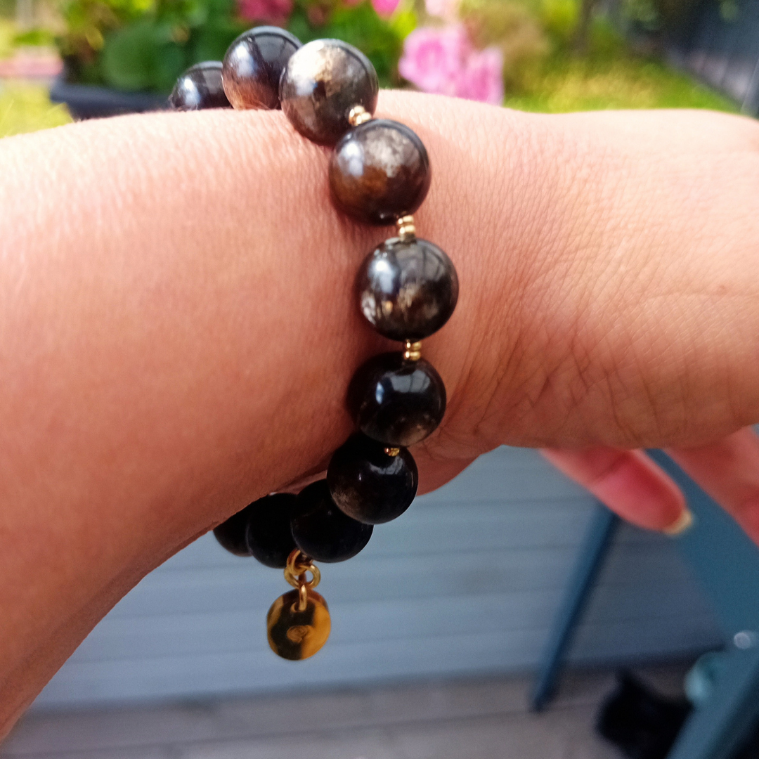 UMBRA-armband på handleden med mörka runda golden mica-pärlor, små guldfärgade distanser och en rund guldberlock som logohänge från HerMine`s